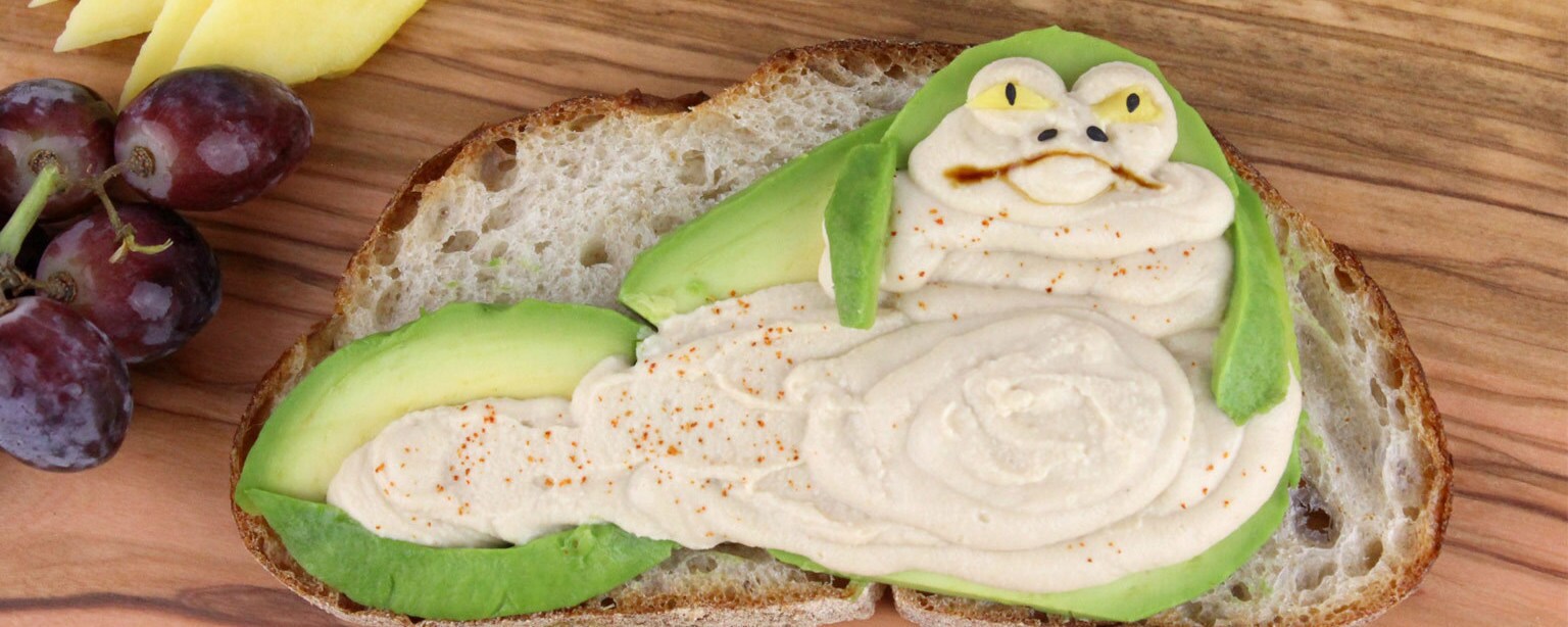A slice of bread covered with avocado and hummus in the shape of Jabba the Hutt with fruit on the side.