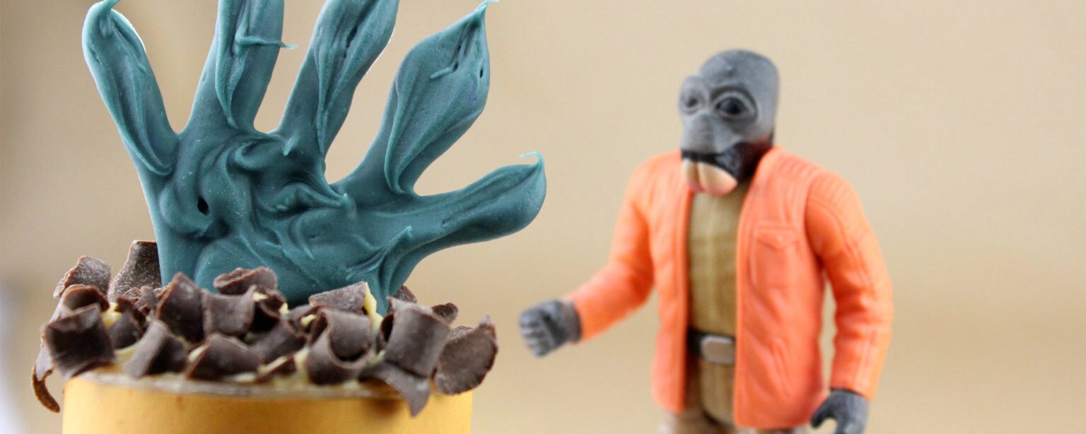 A yellow push pop topped with green chocolate molded into Ponda Baba's hands next to a Ponda Baba action figure.