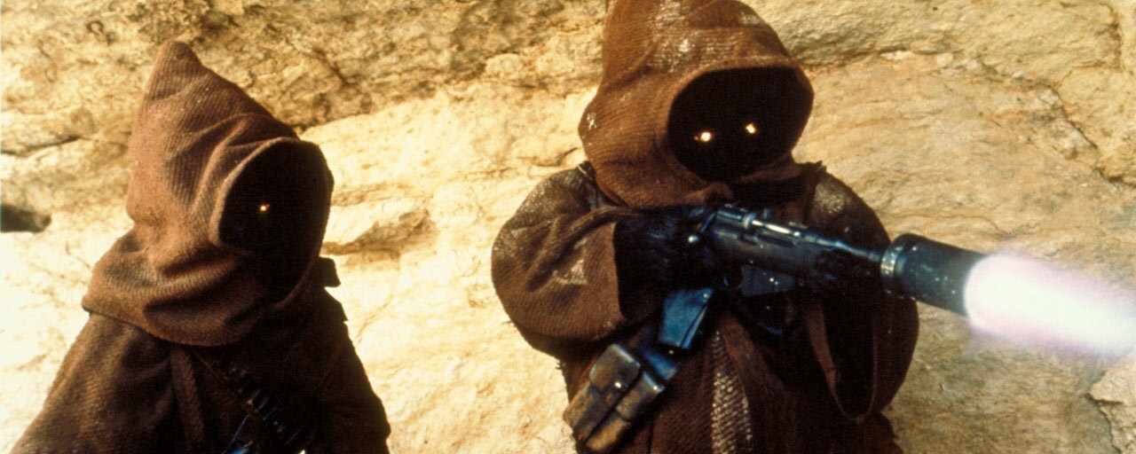 Two Jawa scavengers stand in front of a large desert rock as one fires an ion blaster weapon, their glowing eyes visible beneath their hoods.