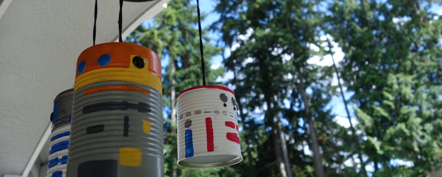 A completed droid wind chime hangs from an overhang with trees looming in the background.