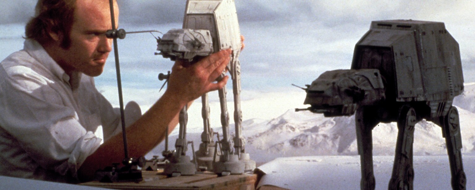 A stop-motion artist positions a model AT-AT in front of a Hoth backdrop while a second AT-AT stands close by in a behind-the-scenes image from the making of The Empire Strikes Back.