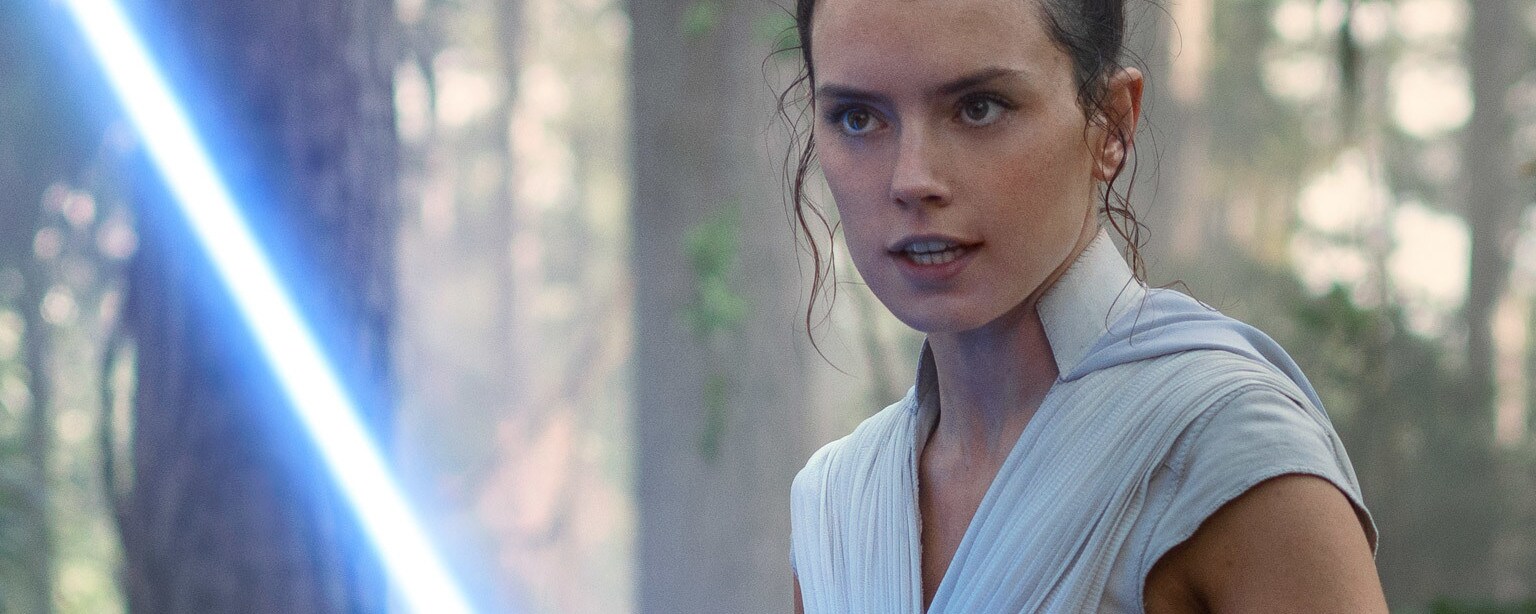 Rey holds her lightsaber in a forest.