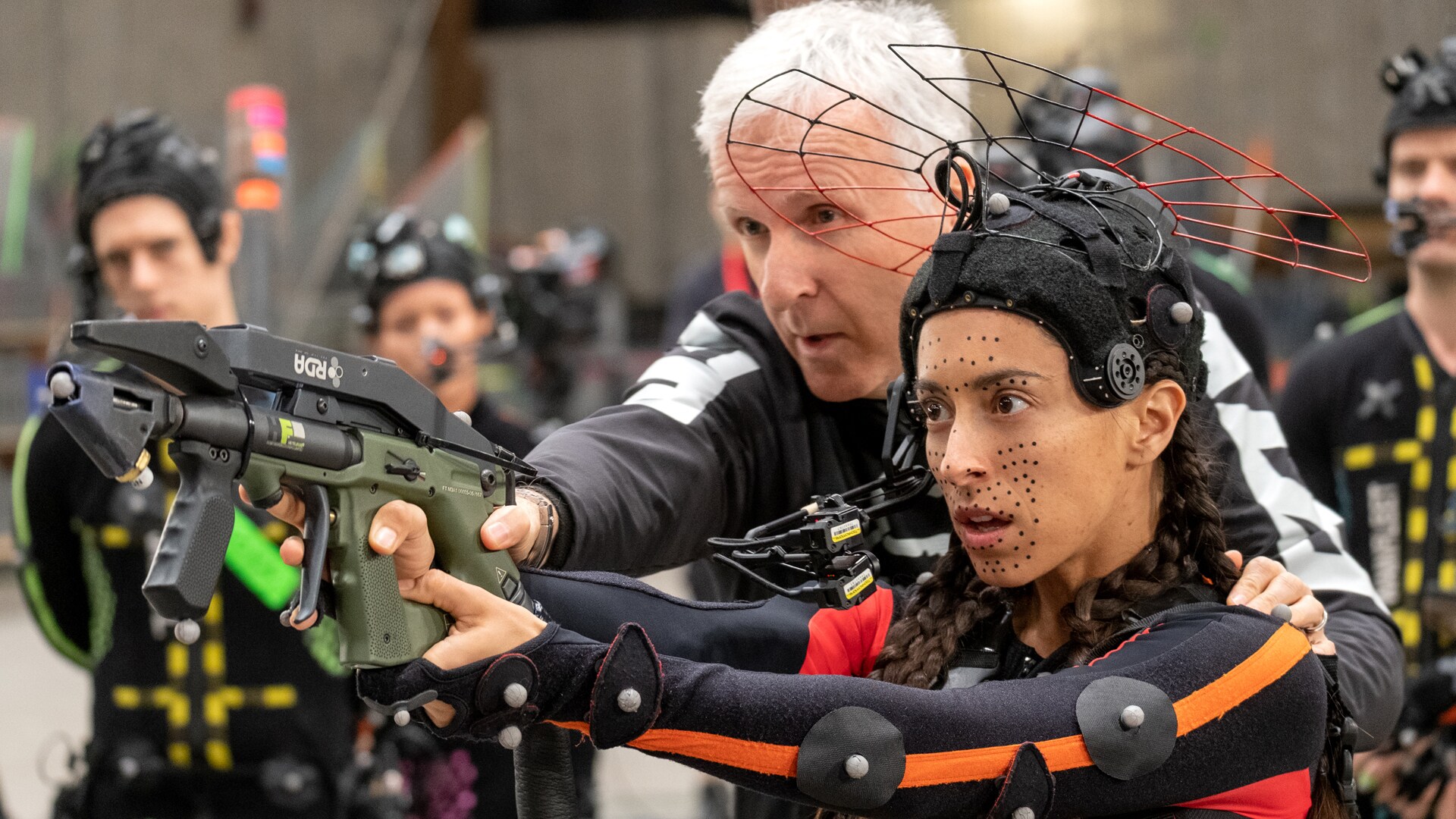 Image of Director James Cameron and actor Oona Chaplin. Cameron is assisting Chaplin, who is in a motion capture suit, with the aiming of a prop gun. There are other actors in the background observing.