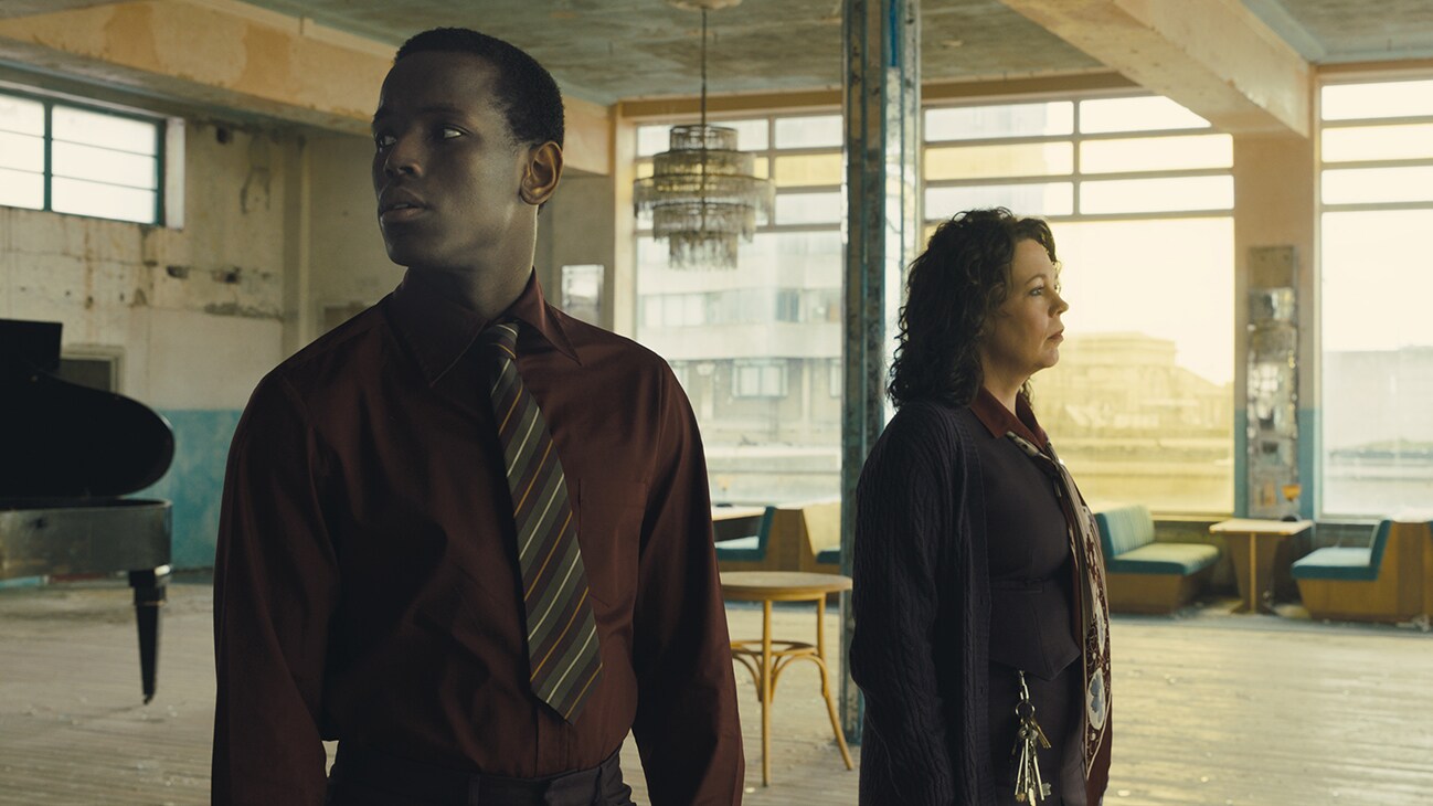 Olivia Colman and Michael Ward stand in an empty and dusty and decrepit room appearing to look concerned. The sun shines through floor to roof windows in the background, a lone pigeon sits on the floor, with a grand piano sitting further in the background.