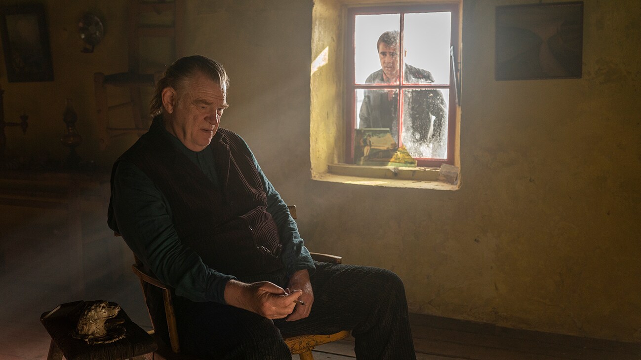 Brendan Gleeson's character sits on a chair within a dark room, gripping a cigarette and looking towards the floor, Colin Farrell's character peers in through a dirty but sunny window - a confused look on his face.
