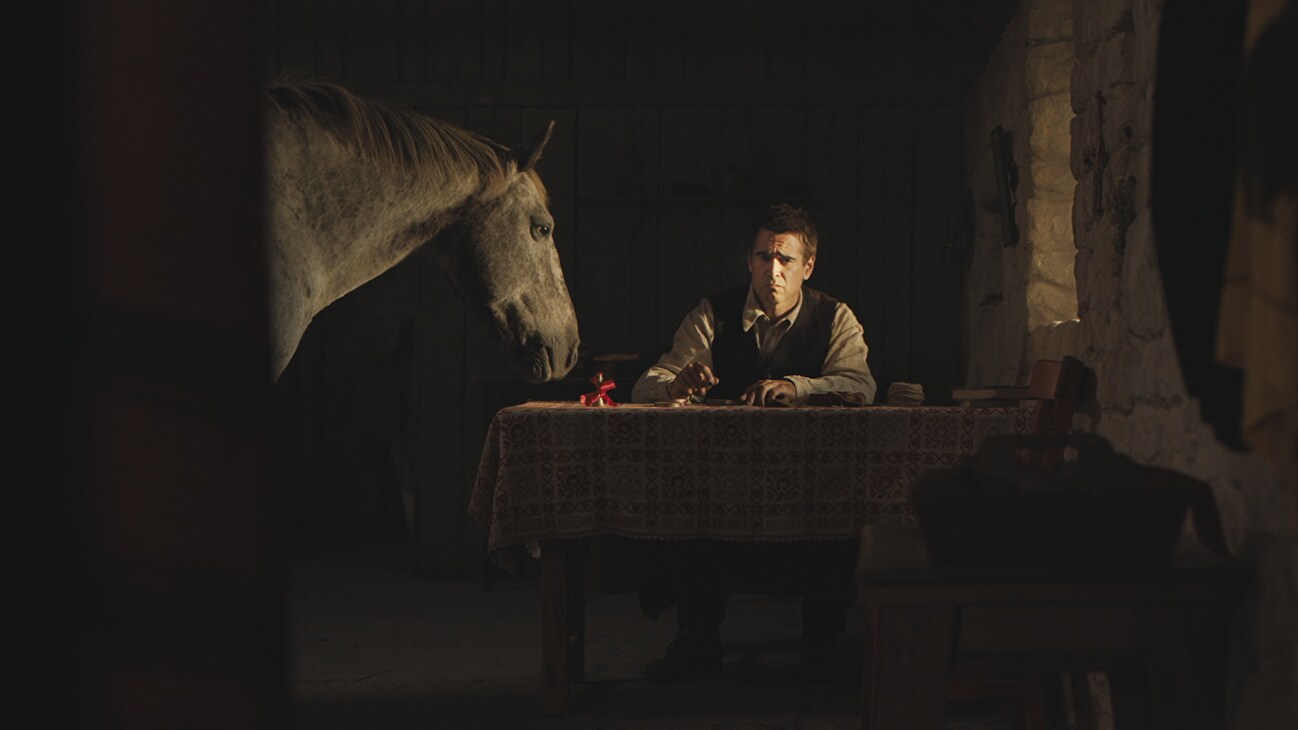 Colin Farrell's character sits at a dinner table inside a dark room, a horse peers into the room from the left of the image, it's head sitting calmly above the table.