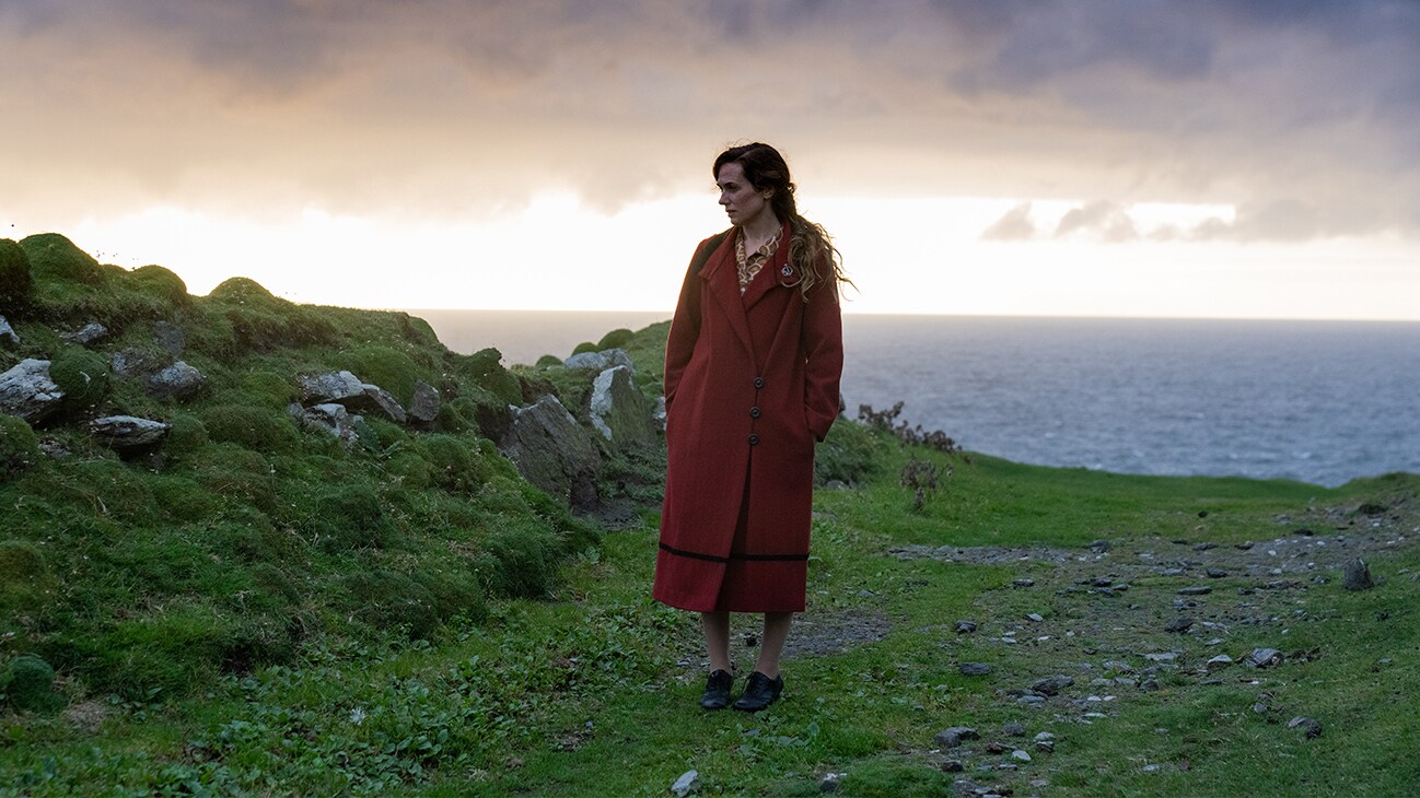 Kerry Condon's character stands in a green outcrop, overlooking the ocean, looking towards the ground, wearing a long red dress.