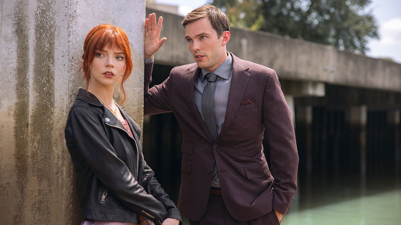 Anya Taylor-Joy looks away in a black leather jacket, clutching a purse, while Nicholas Hoult looks towards her in a dark maroon suit, appearing to speak. The pair are stood outside in the sunshine.