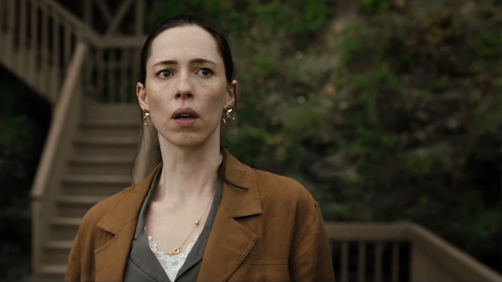 Actor Rebecca Hall with a scared expression standing in front of an outdoor staircase in the movie The Night House