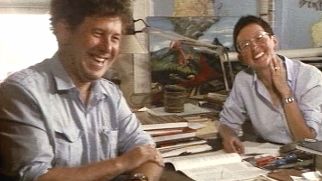 Katia and Maurice Krafft sit in their office surrounded by books, papers and images of volcanoes, laughing at the camera while being interviewed.