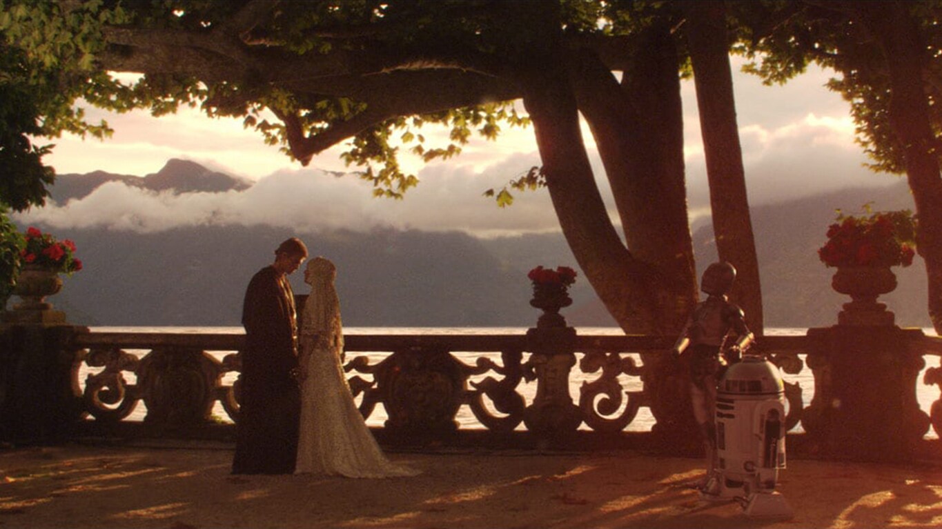 Anakin Skywalker and Padmé Amidala stand holding each others hands closely at Varykino lake retreat in Naboo's Lake Country, droids R2-D2 and C-3PO stand to the side witnessing their marriage.