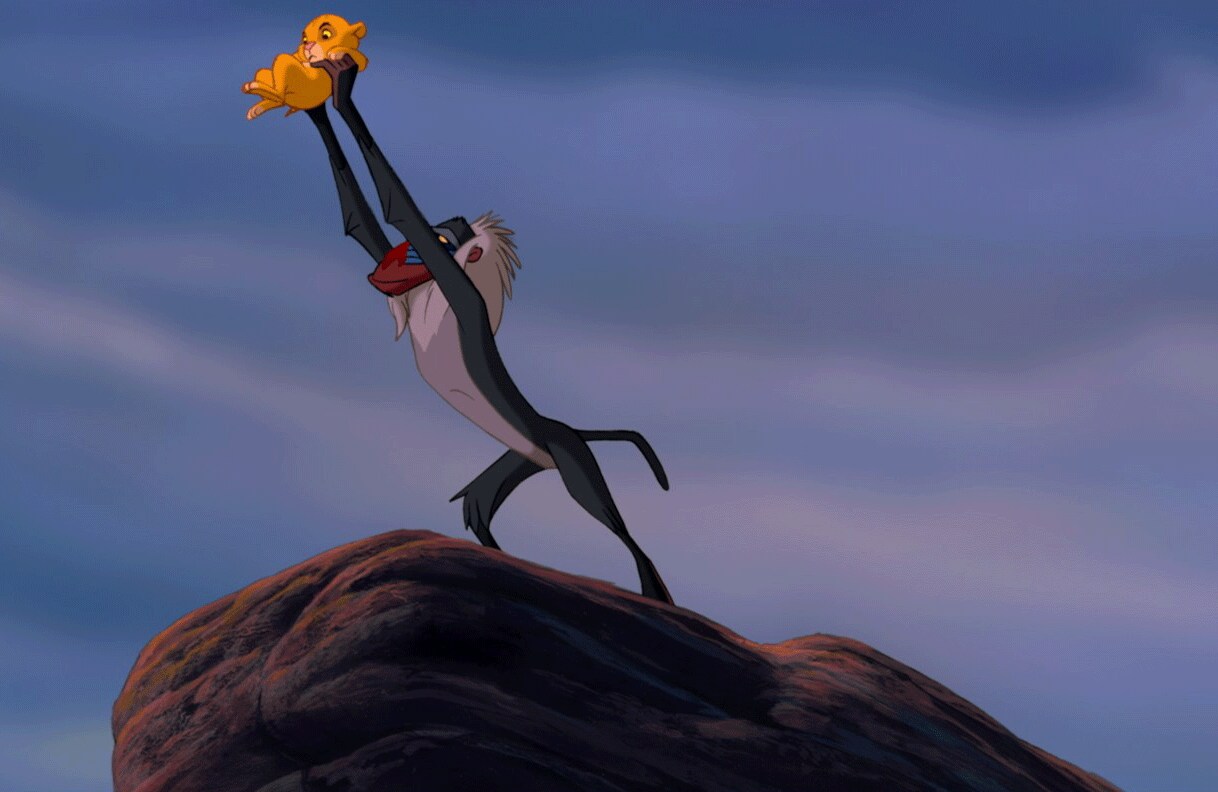 A still from Disney's The Lion King movie showing Rafiki holding baby Simba above his head as he stands atop Pride Rock.