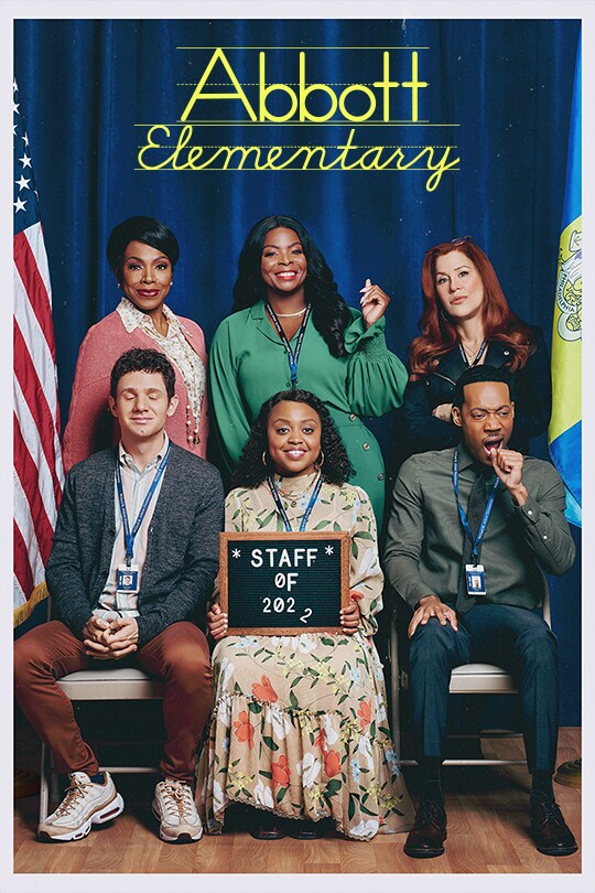 The mixed cast of characters from Abbott Elementary sit and stand while having a staff photo taken in front of a dark blue draped cloth. 
