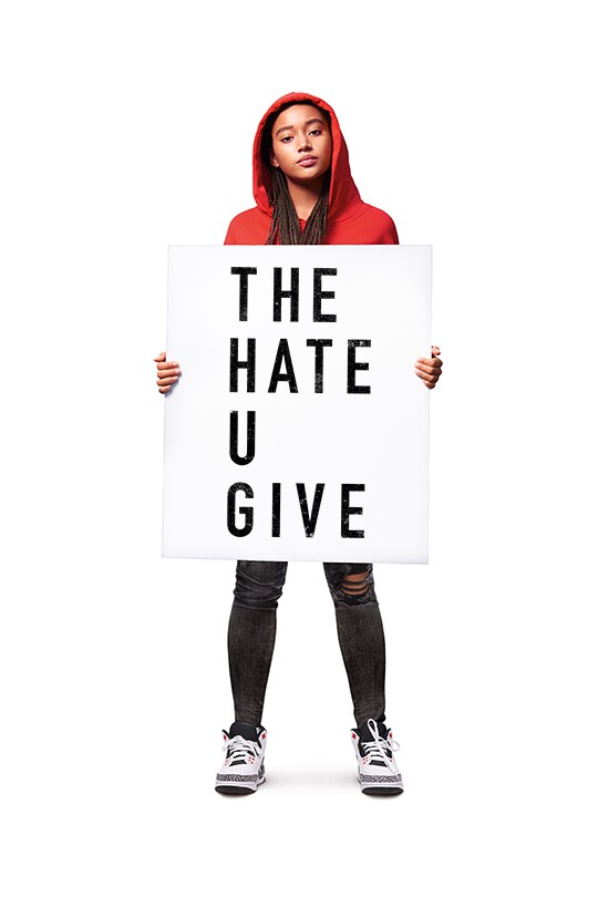 Amandla Stenberg as Starr Carter stands in a red hoodie and black pants, holding a white and black sign which says 'The Hate U Give'