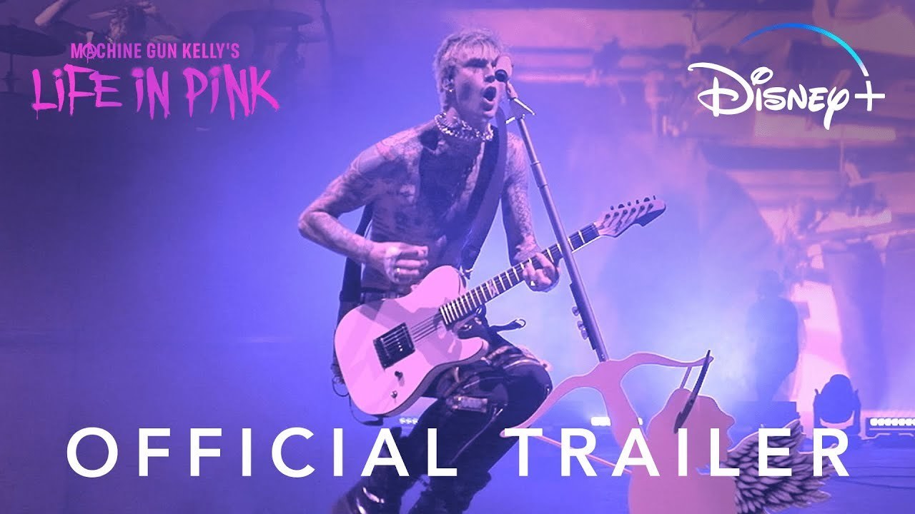 Musician Machine Gun Kelly is mid-air, holding his white guitar in front of a microphone. The image has a pink-hue to it, the title 'Machine Gun Kelly: Life In Pink' is in the top left hand of the image, the Disney+ logo in the top right, and 'Official Trailer' at the bottom.
