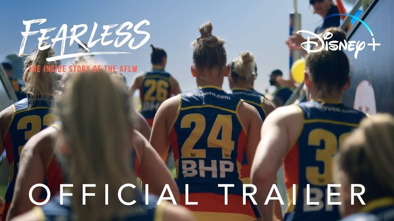 AFLW players head out of the dressing sheds towards the field through a tunnel, fans are on either side cheering them on. The 'Fearless: The Inside Story of the AFLW' title is in the top left hand side, 'Disney+' in the top right and 'Official Trailer' below.