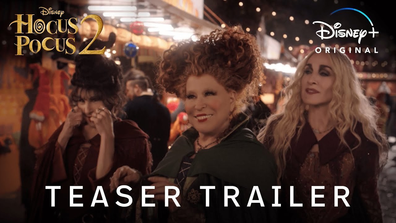 The three witches from Hocus Pocus 2 look towards an unknown person, they appear to be walking down a path with a carnival in the background.