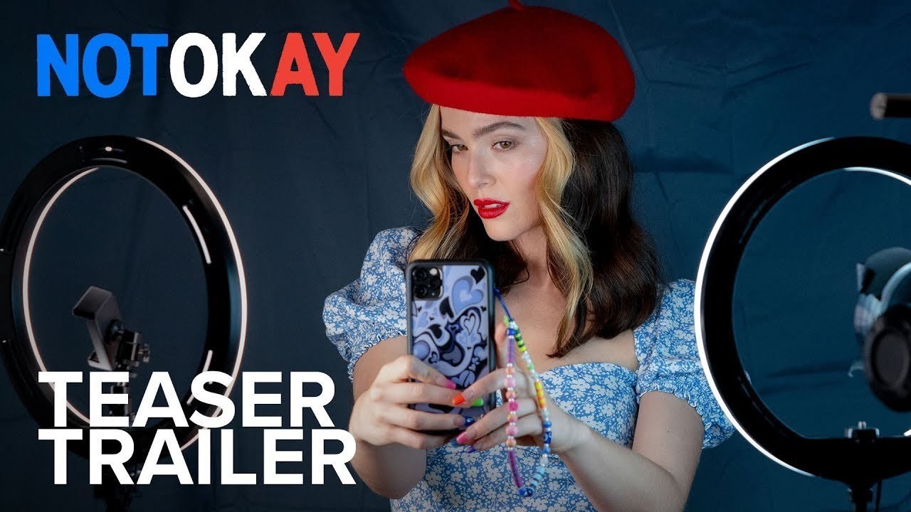 Zoey Deutch looks at her phone, appearing to be taking a selfie with two ring lights brightening her face, she is wearing a blue dress with flowers and wearing a red beret style hat. The 'Not Okay' title treatment is in the top left hand corner of the image.