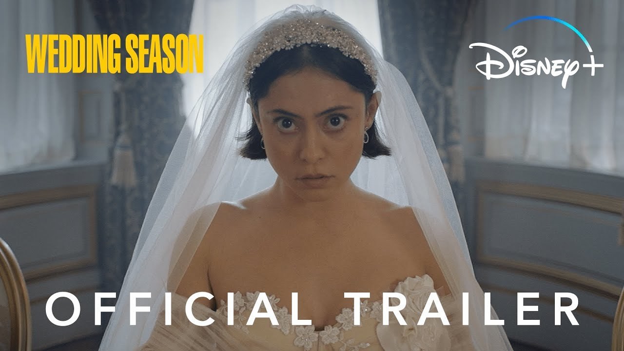Rosa Salazar looks towards the camera with a blank expression, wearing a wedding dress and veil, with a blue wall and curtains in the background. The 'Wedding Season' title is in the top left hand of the image, 'Disney+' in the top right and 'Official Trailer' on the bottom.