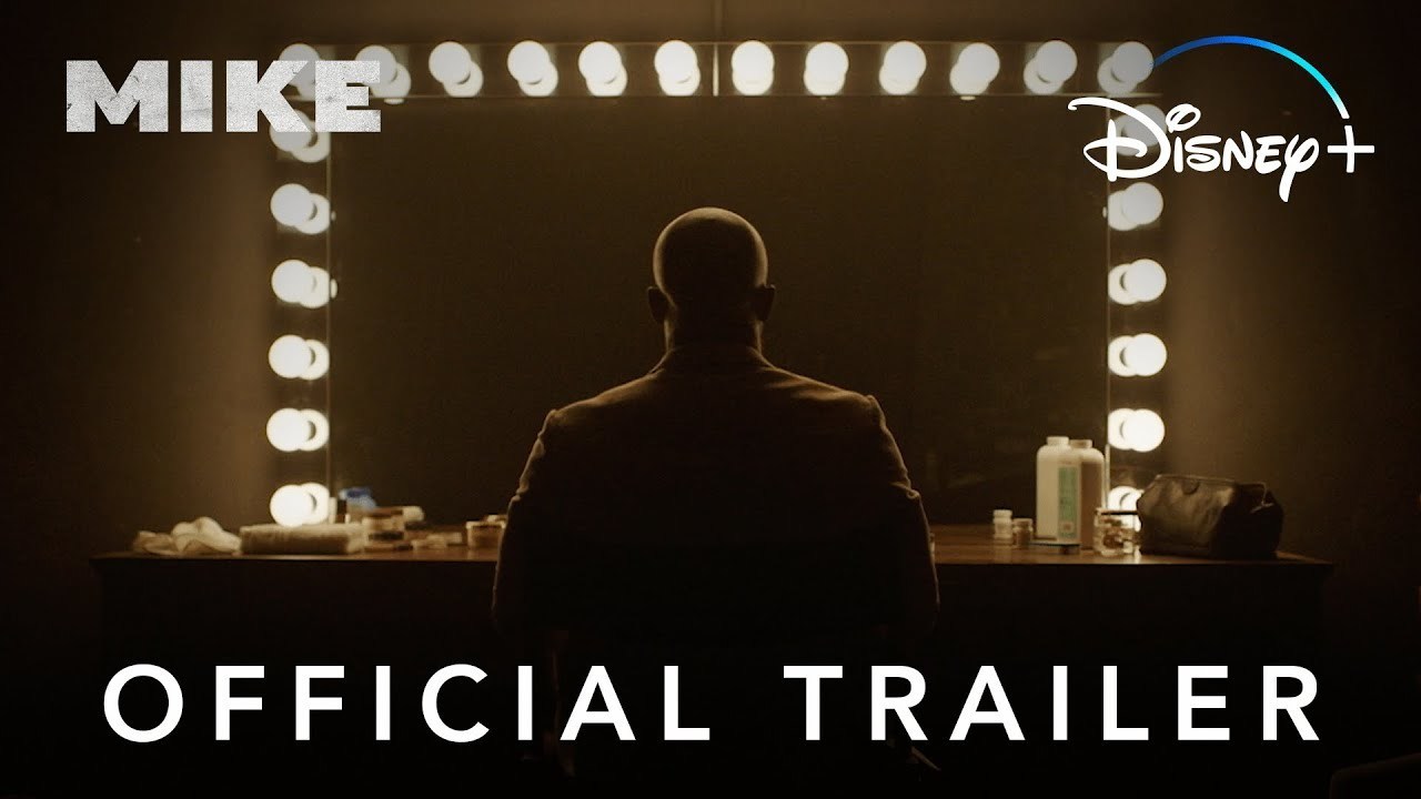 An actor playing former professional boxer Mike Tyson looks into a Hollywood style large vanity mirror, the image only shows the silhouette of his back. The title of the series 'Mike' is in the top left hand of the image, 'Disney+' is in the right while 'Official Trailer' is in the foreground.