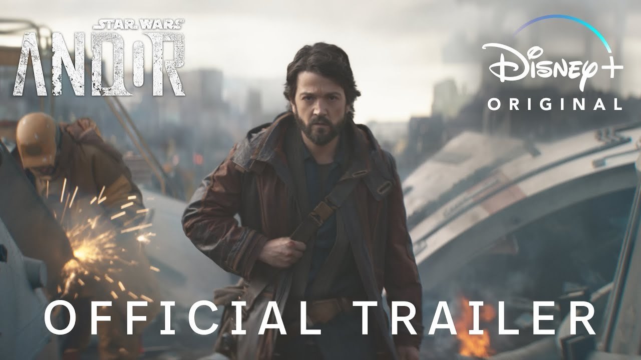 Diego Luna as Cassian Andor walks towards the camera in a dark brown coat, smoke billows in the background as two people weld metalwork.