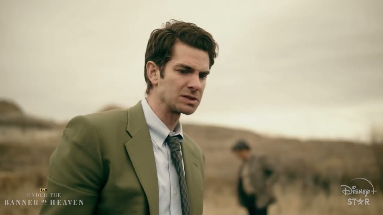 Andrew Garfield stands looking bothered in a overcast field, shot from the chest up, with an unknown character out of focus in the background.
