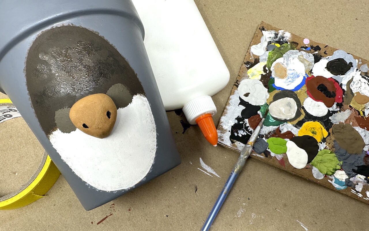 Next, use the black acrylic paint to paint the nostrils of Bazil’s nose and his two eyes. Let dry. Use the end of a paint brush to add two white dots on the eyes as reflections. Next, paint his nose with the tan acrylic paint and let dry.