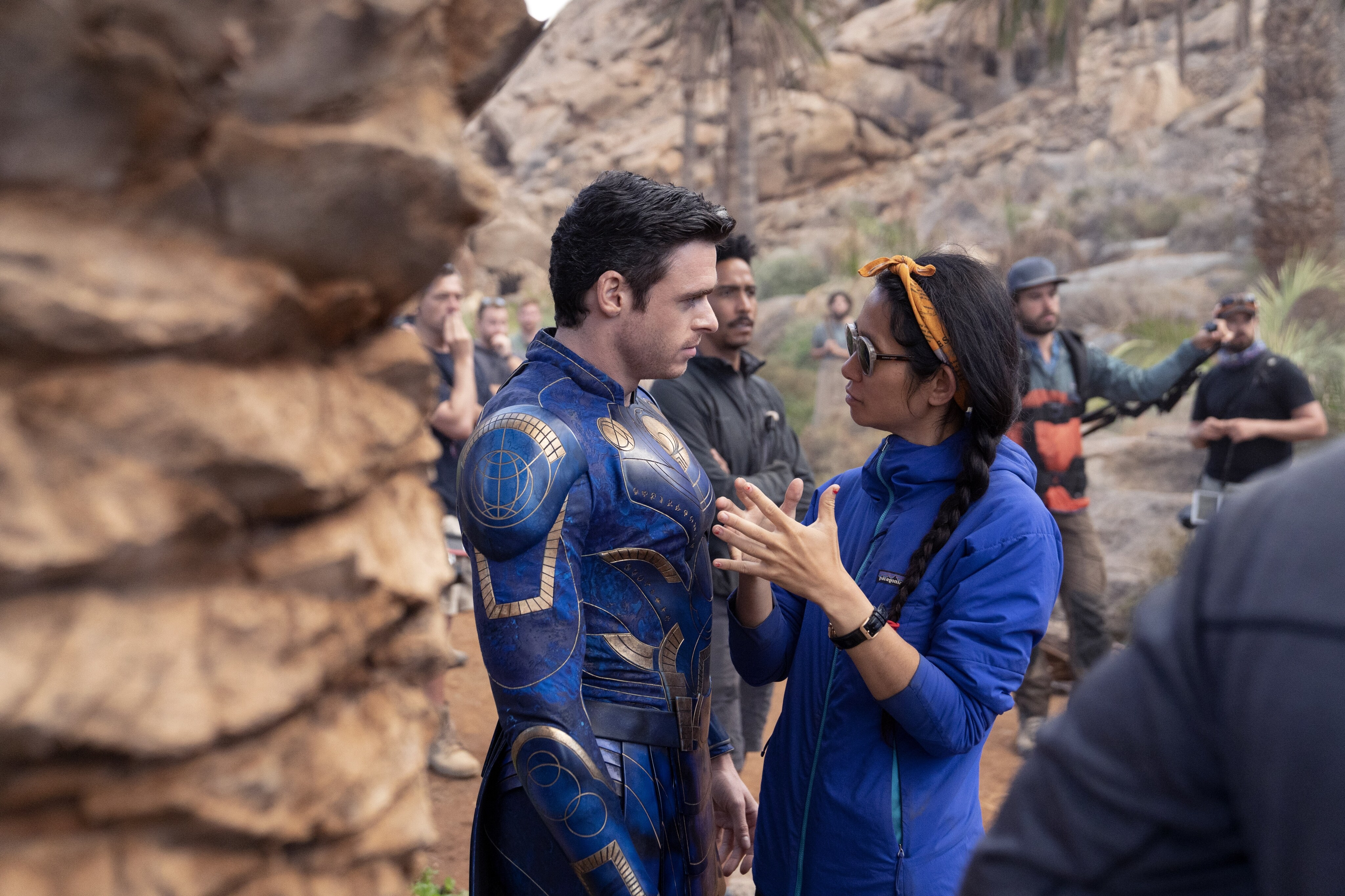 Chloé Zhao directs Richard Madden as Ikaris in Marvel Studios' Eternals. The background is a desert canyon and there are other crew members visible in the background. Madden wears a supersuit.