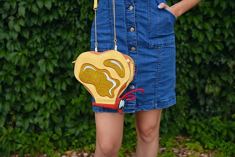 A close-up of a woman's purse that looks like a butterfly composed of pieces of toast with butter on it. The woman is wearing a denim overall dress