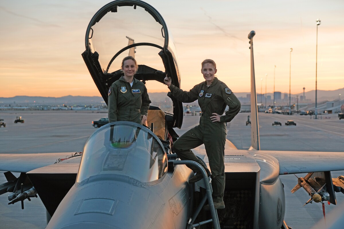 Brie Larson as Carol Danvers stands on a fighter plane with another woman, as the sun sets in the background in a scene from Marvel Studios' Captain Marvel.