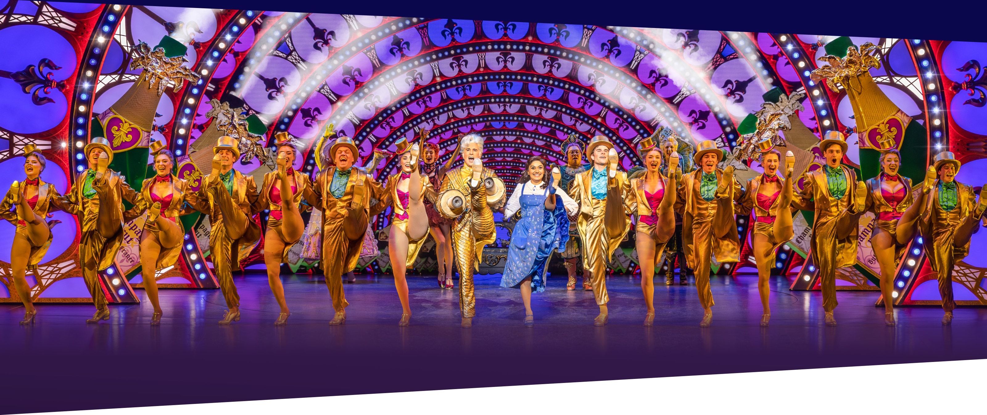 Belle und Lumière lächeln sich an und tanzen zusammen mit dem Ensemble. Die Darsteller:innen stehen in einer Reihe und schwingen ihr Bein in die Luft. Sie tragen goldene Anzüge. Mittig steht Belle in einem blauen Kleid.