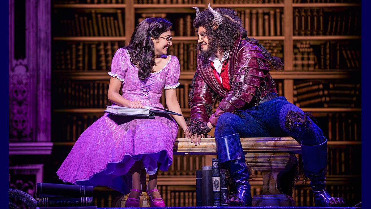 Belle sitzt gemeinsam mit dem Biest auf einer Bank. Ihre Hände berühren sich und die beiden schauen sich freudig in die Augen.