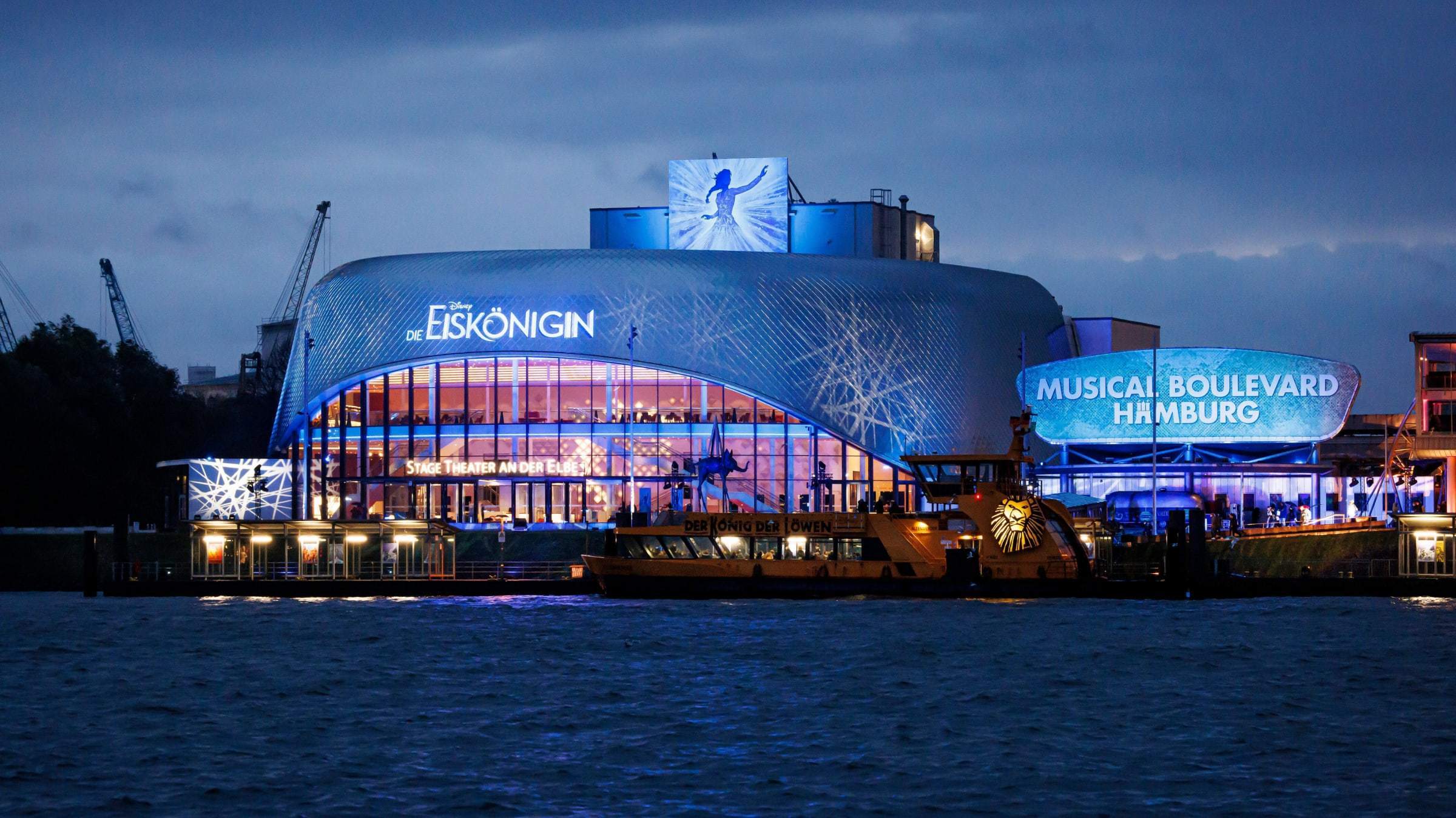 Das Stage Theater an der Elbe in Hamburg mit dem Schriftzug "Disneys Die Eiskönigin", wie es in der Dämmerung in blaues Licht getaucht wird.
