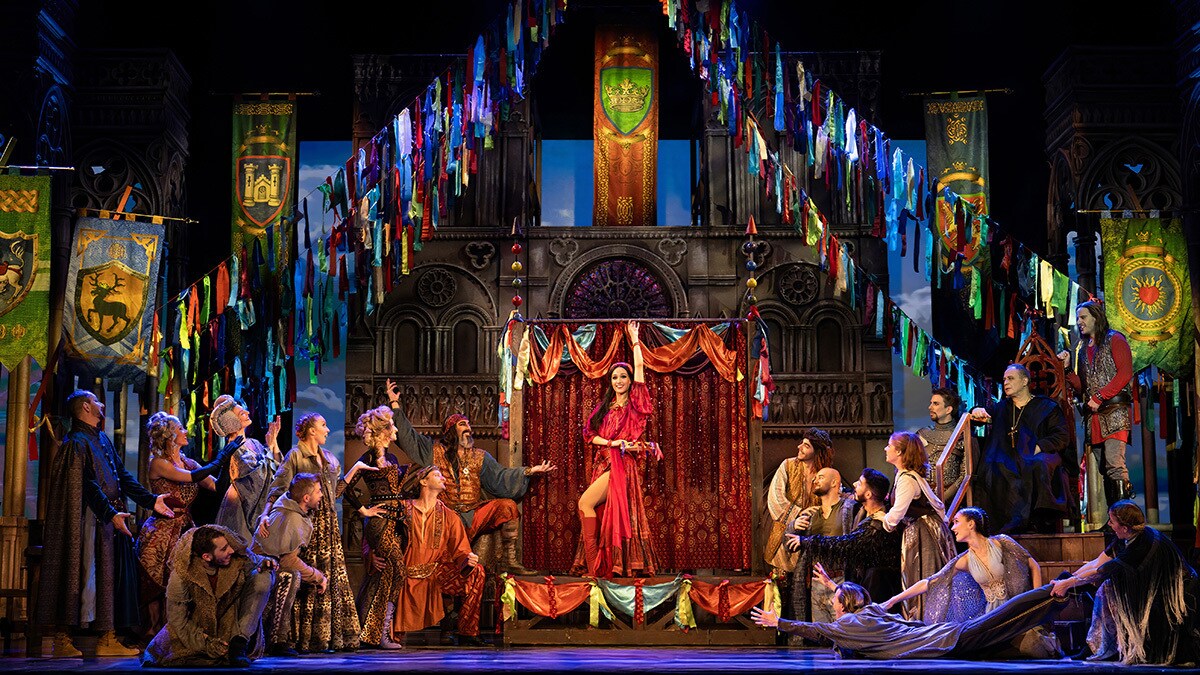 Esmeralda tanz beim Narrenfest auf der Bühne und reißt die Aufmerksamkeit an sich. Viele Menschen sind versammelt und schauen interessiert zu ihr. 