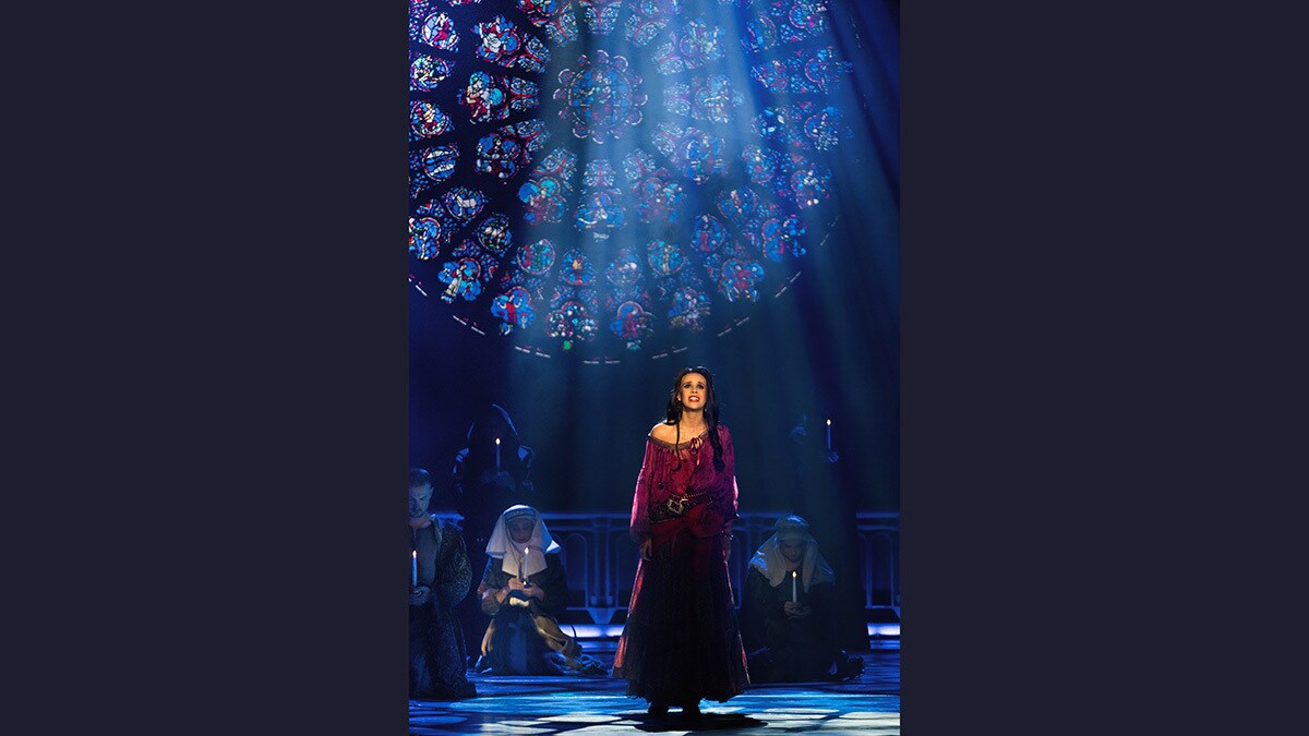 Esmeralda steht vor einem schmuckvollen und bunten Kirchenfenster. Im Hintergrund sind Menschen, die Kerzen halten.