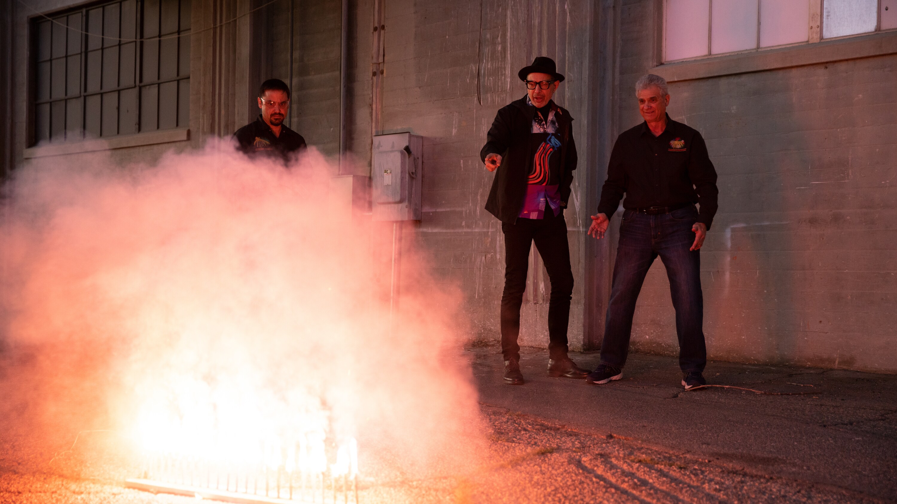 San Bernardino, CA - Jeff Goldblum (center) and Chris Souza (R) watch a small ground level fireworks display being controlled by a Pyro Spectacular employee (L). (Credit: National Geographic/Justin Koenen)