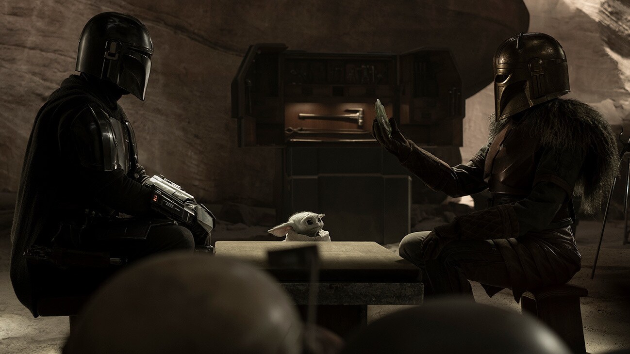 Din Djarin (Pedro Pascal), Grogu and the Armorer (Emily Swallow) sit at a table in the Disney+ Original series, Star Wars: The Mandalorian, Season 3.