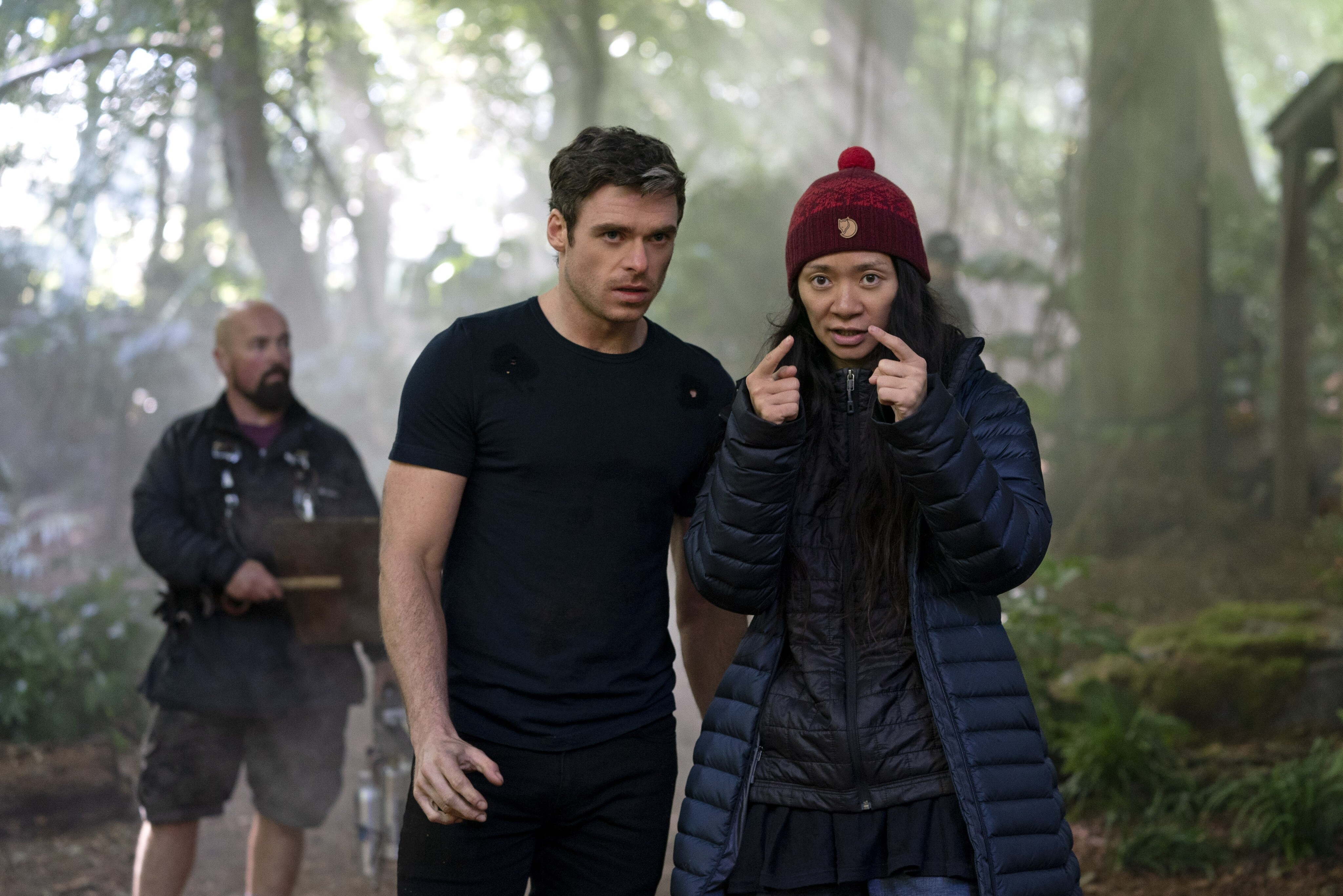 Richard Madden listens as Chloé Zhao directs in a forest setting. A crew member is visible in the background. Madden wears a black t-shirt and black jeans.