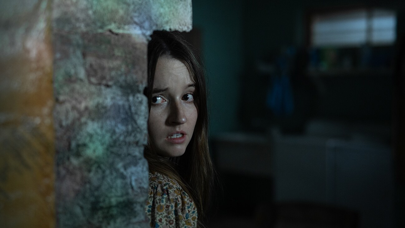 Actor Kaitlyn Dever hiding behind a wall in the 20th Century Studios movie, "No One Will Save You."