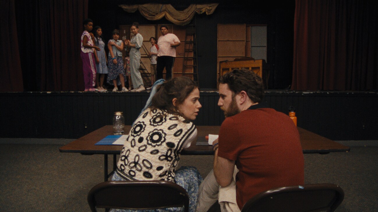 Molly Gordon and Ben Platt sitting at a table talking to each other from the Searchlight Pictures movie, "Theater Camp."