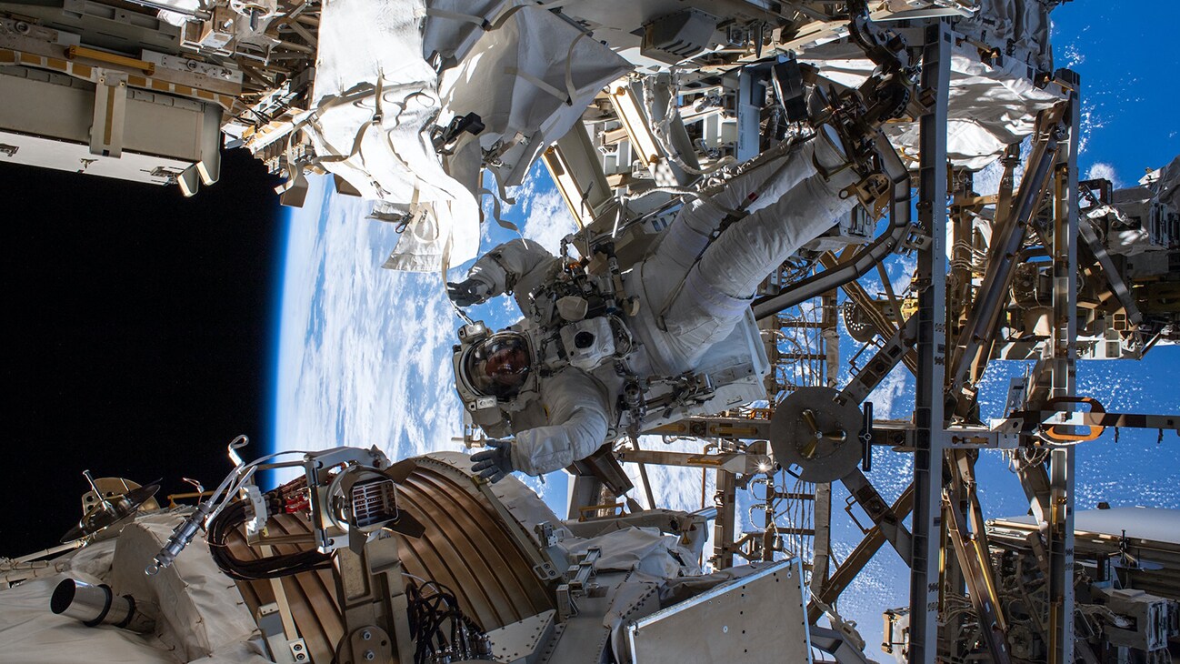 AMONG THE STARS - (Nov. 15, 2019) - NASA astronaut Andrew Morgan waves as he is photographed seemingly camouflaged among the Alpha Magnetic Spectrometer (lower left) and other International Space Station hardware during the first spacewalk to repair the cosmic particle detector. (NASA) ANDREW MORGAN