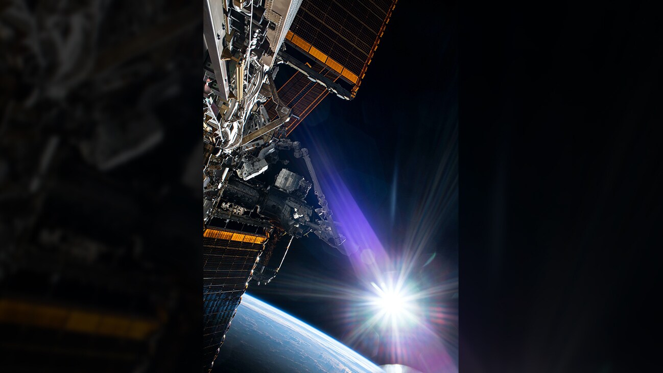 AMONG THE STARS - (July 21, 2020) - The sun beams just above the Earth's horizon as NASA astronaut and Expedition 63 Commander Chris Cassidy (center left) conducts his fourth spacewalk this year at the International Space Station. Cassidy has completed 10 spacewalks throughout his career for a total of 54 hours and 51 minutes spacewalking time. (NASA)  CHRIS CASSIDY