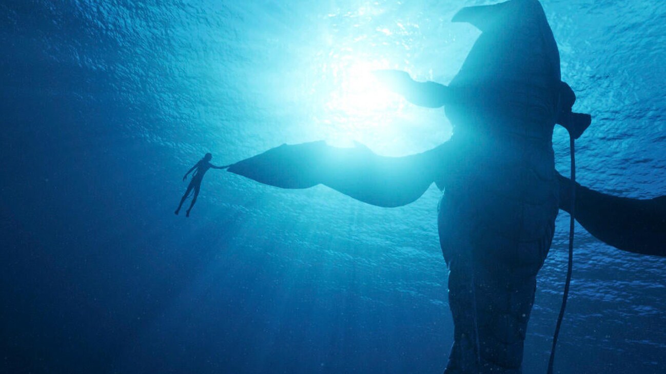 Image of a character holding a fin of a large sea creature underwater from the 20th Century Studios movie Avatar: The Way of Water.