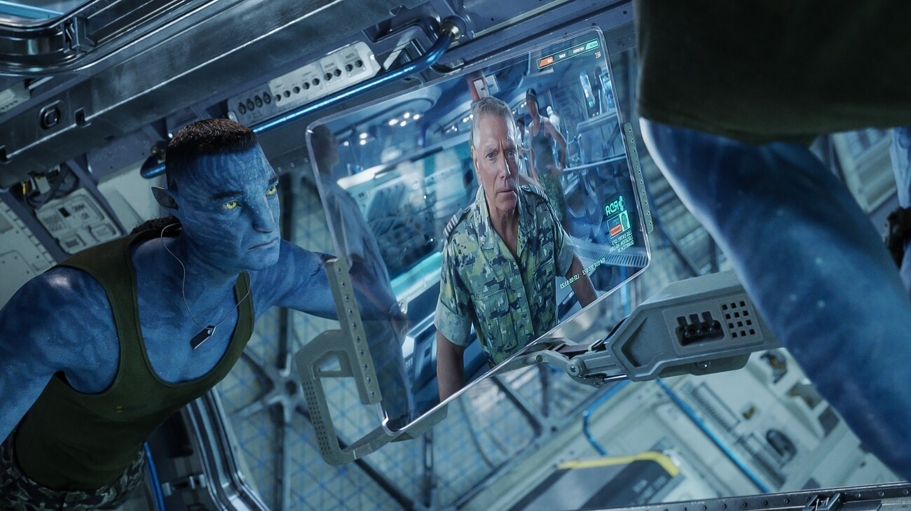 A Na'vi dressed in combat gear looks on as Colonel Quaritch appears on a screen.