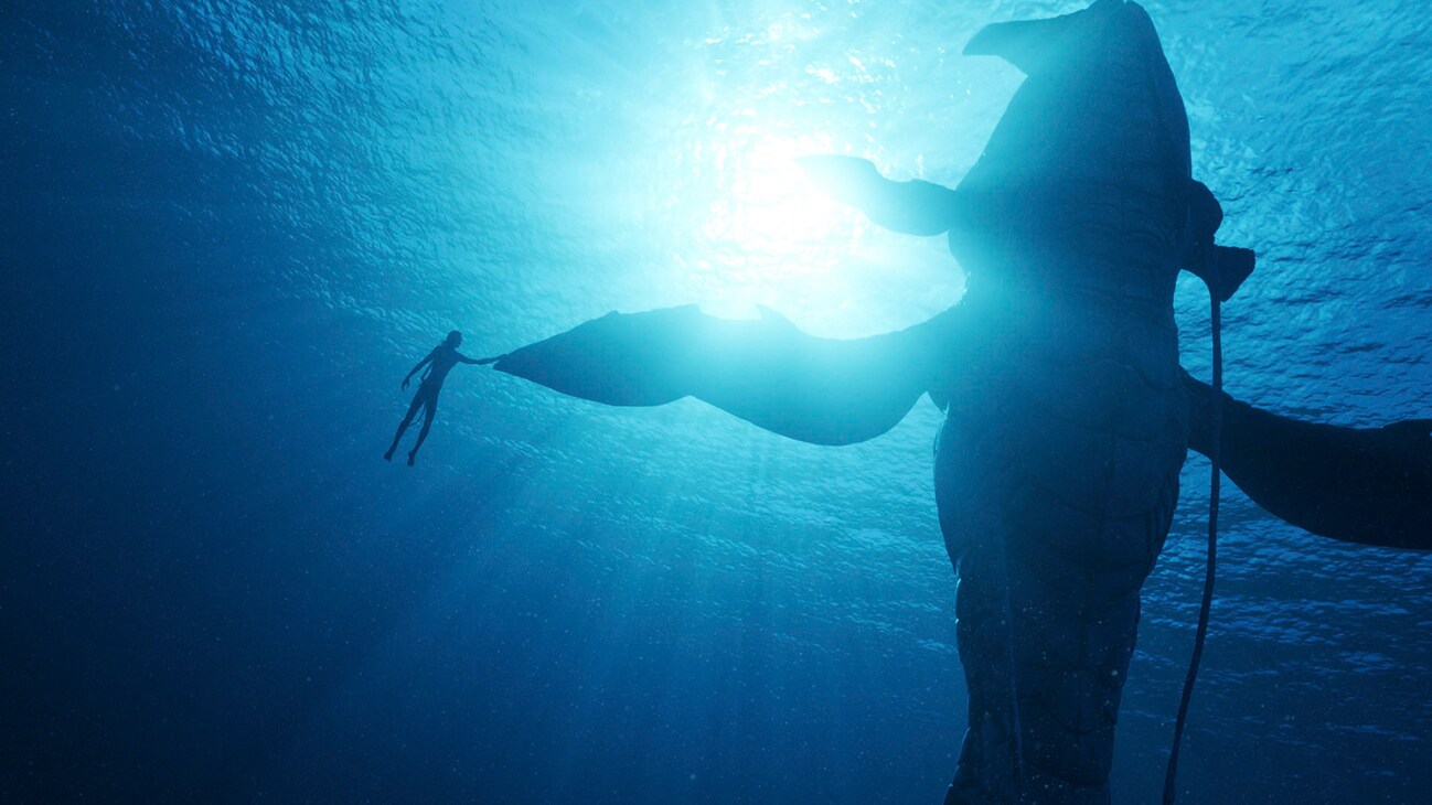 Image of a character under water swimming by a large sea creature from the 20th Century Studios movie Avatar: The Way of Water.