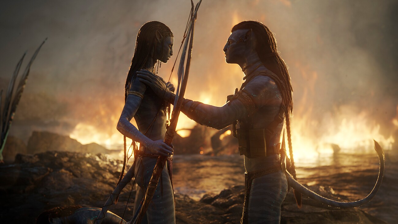 A Na'vi male and female staring at each other as fire burns in the background from the 20th Century Studios movie, "Avatar: The Way of Water".