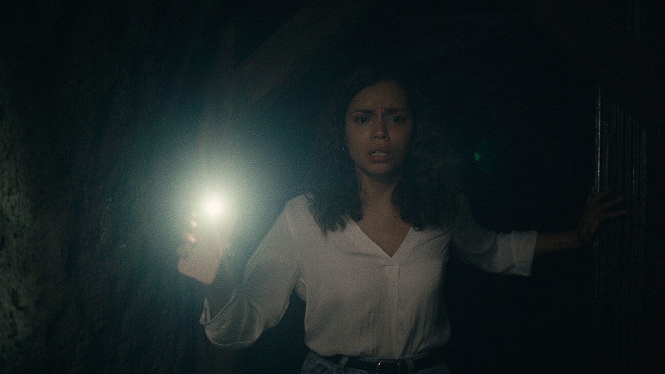 A young woman (actor Georgina Campbell) in a dark room holding a cell phone in front of her with the light turned on from the 20th Century Studios movie "Barbarian".