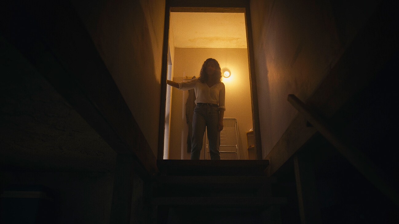 A young woman (actor Georgina Campbell) standing at an open door peering down a dark staircase from the 20th Century Studios movie "Barbarian".