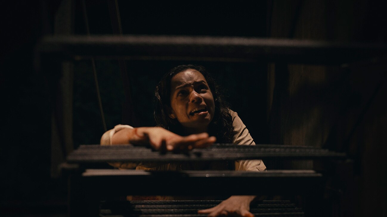 A young woman (actor Georgina Campbell) with a terrified look crawling up a dark staircase from the 20th Century Studios movie "Barbarian".