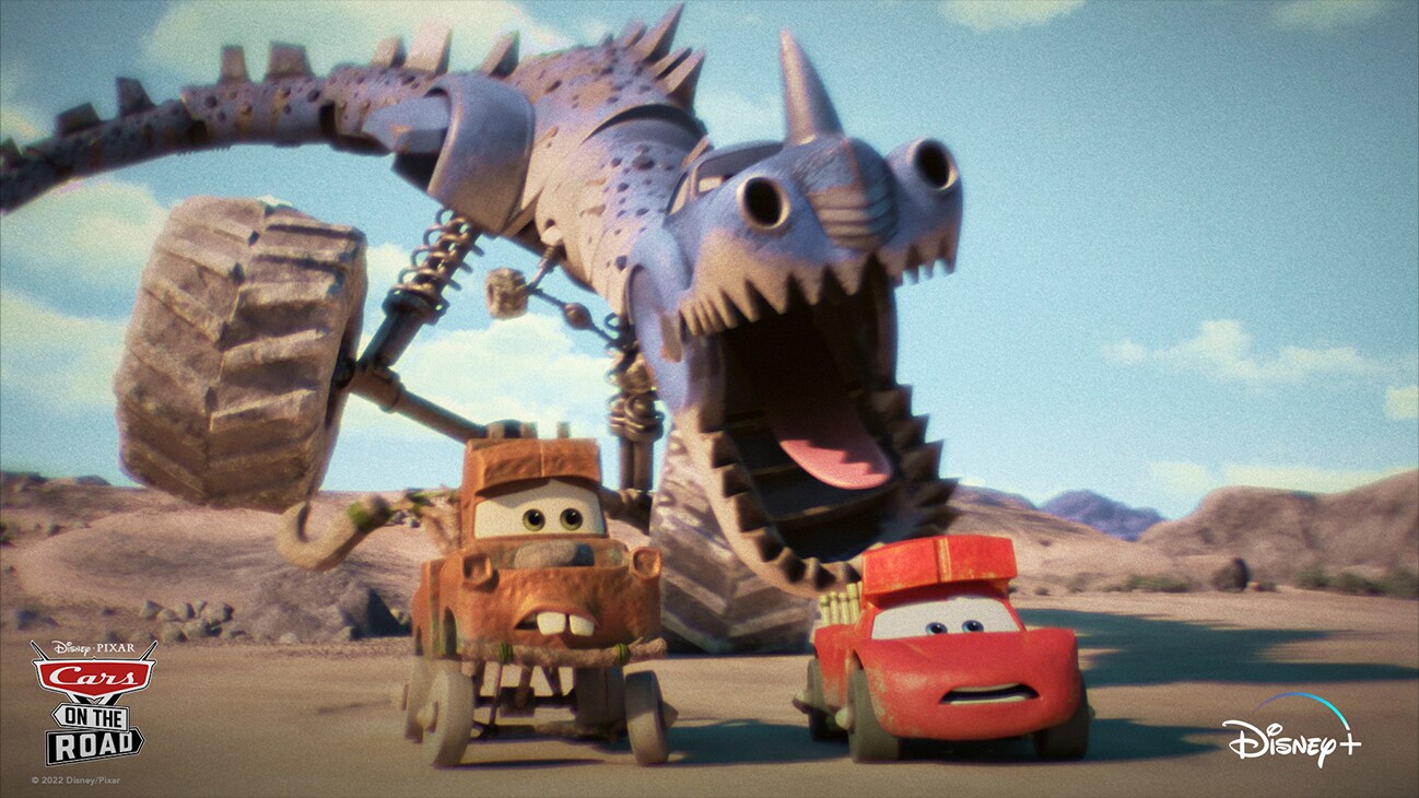 Pre-historic Lightning McQueen (voice of Owen Wilson) and Mater (voice of Larry the Cable Guy) being chased on an open plain by a mechanical dinosaur with large wheels from the Disney+ Original series "Cars on the Road".